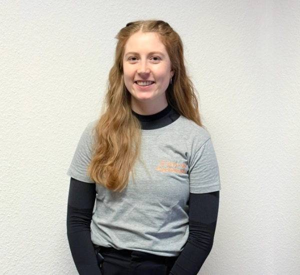 Ergotherapeutin Sabrina Dyylewski steht vor einer weißen Wand in der Praxis. Ihre offenen Haare fallen über ihre rechte Schulter, sie lächelt freundlich und offen in die Kamera.