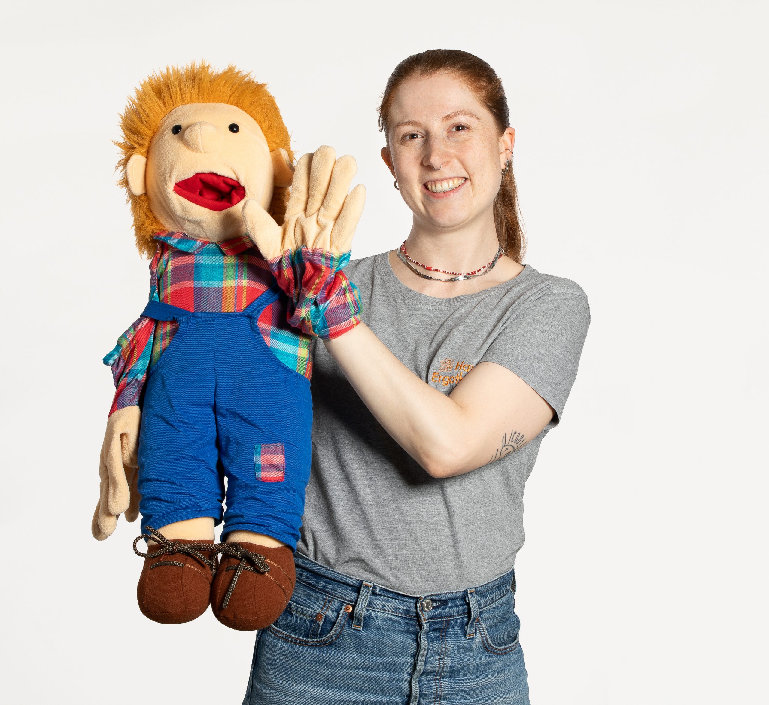 Sabrina Dylewski steht vor einem hellen Hintergrund und lächelt in die Kamera. Sie hält eine bunte Handpuppe mit kariertem Hemd und blauer Latzhose.